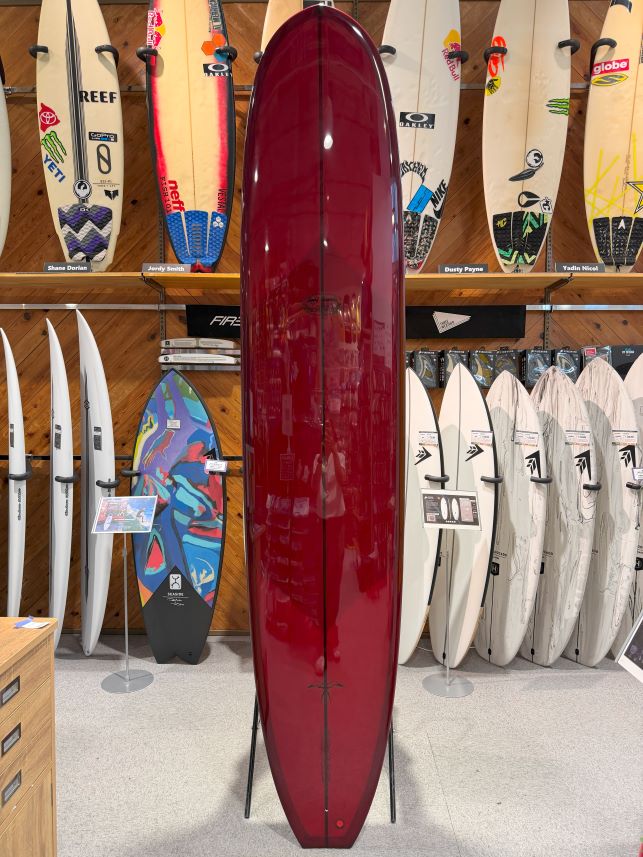Glossy burgundy surfboard on a metal stand in a surf shop, with numerous other boards displayed on racks along the back wall.
