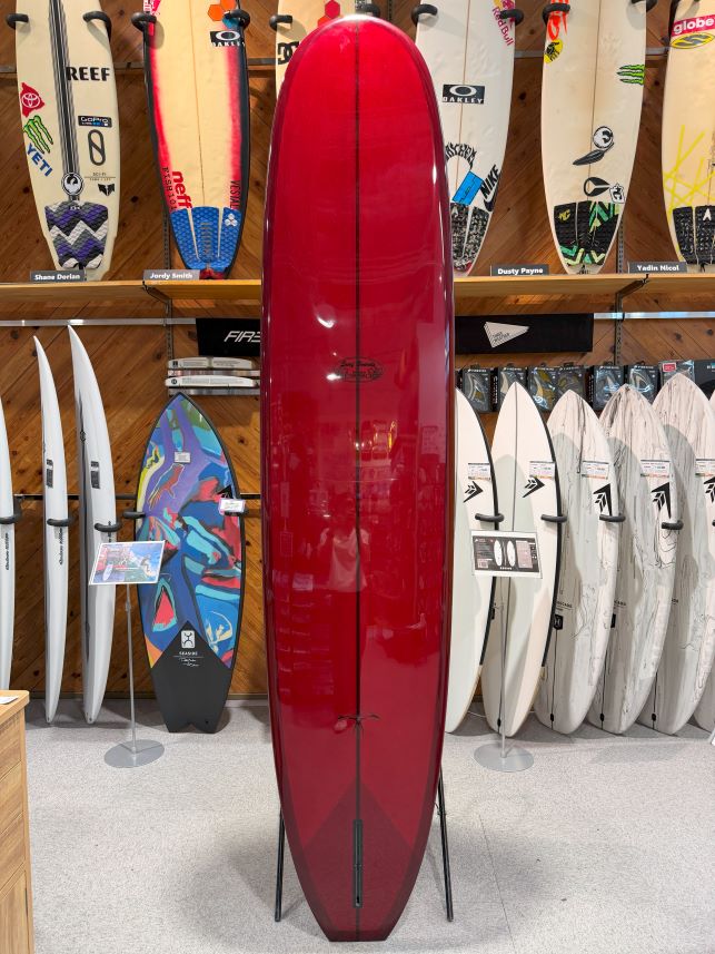 Red longboard surfboard on a stand in a surf shop, surrounded by various white and patterned boards on racks behind it.