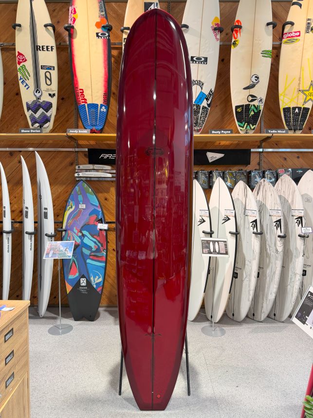 Red glossy surfboard on a stand in a shop display, with numerous boards lined along the walls behind.