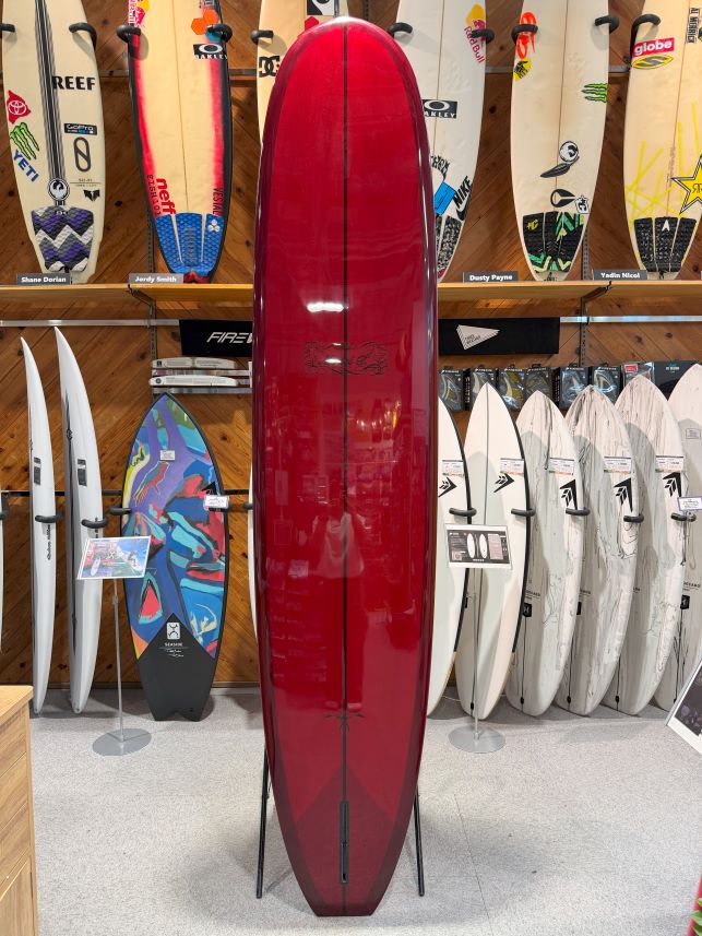 Glossy red surfboard on a stand in a surf shop, surrounded by white and colorful boards on display.