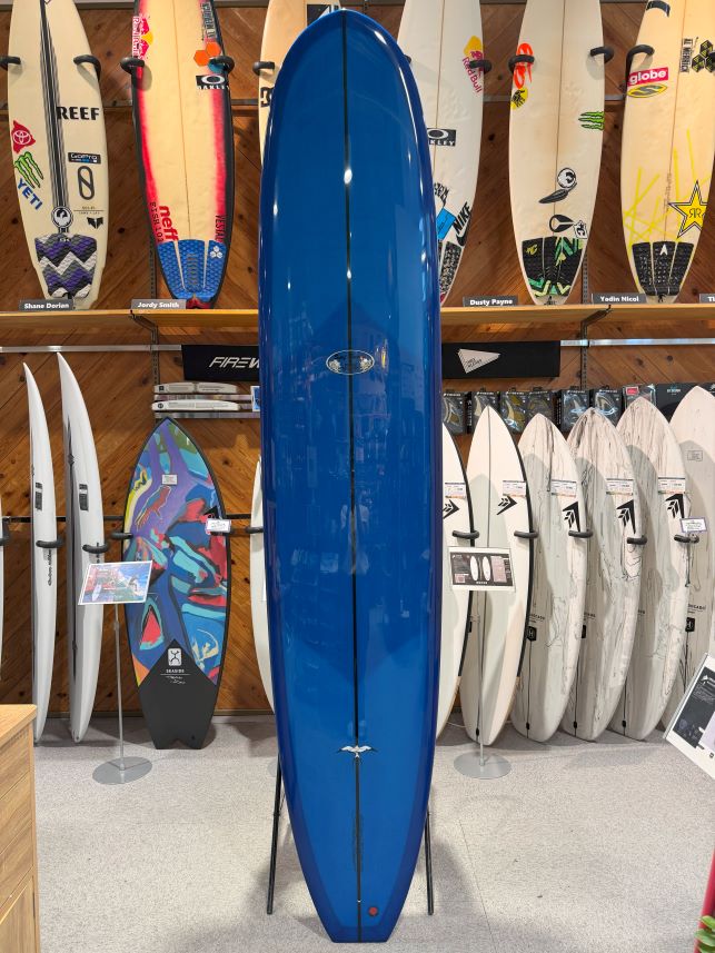 Blue longboard surfboard on a display rack in a surf shop, surrounded by other boards on shelves.