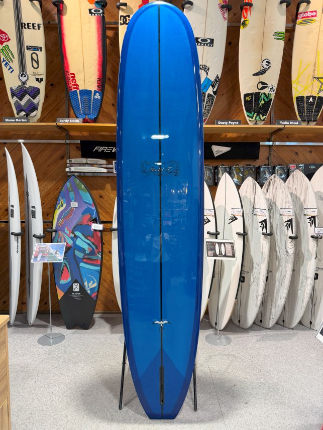 Blue surfboard on a stand in a shop, surrounded by many other surfboards on racks and walls.