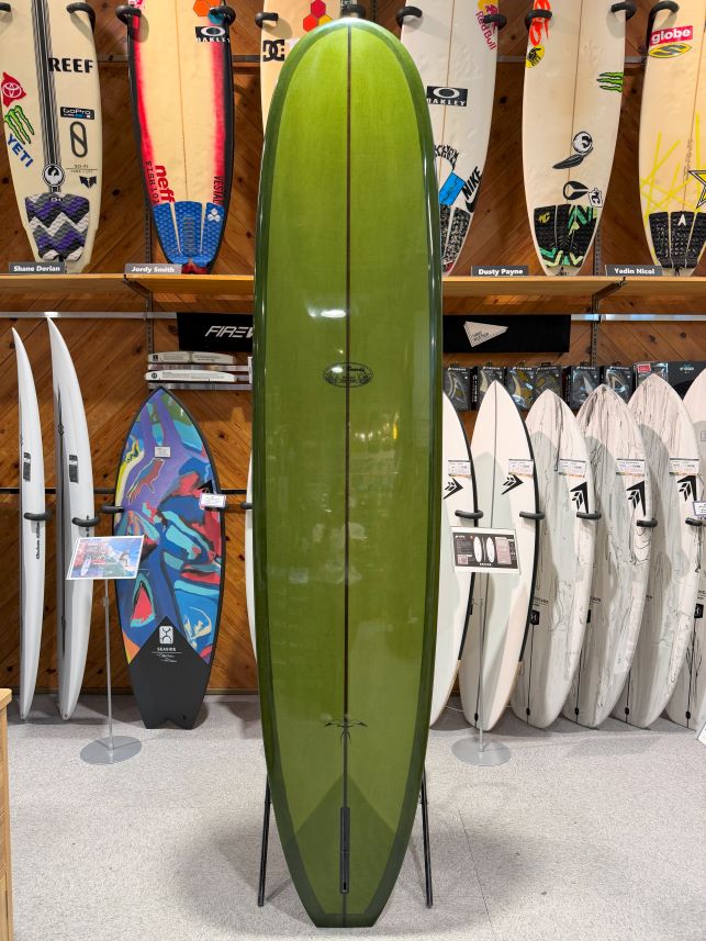Long olive-green surfboard on a stand in a surf shop showroom.