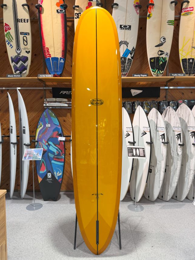 Bright yellow longboard on a display stand in a surf shop, with other boards lining the walls behind it and racks in the foreground.