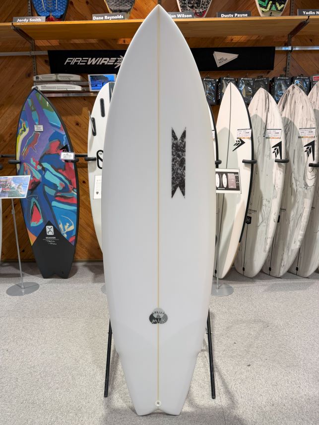 IMG_0305 - SHOP NEWS | ムラサキスポーツオンラインストア 通販 White shortboard surfboard on a display stand in a surf shop, with other boards and gear on shelves behind it.