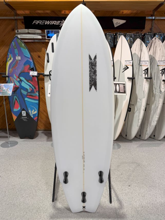 IMG_0306 - SHOP NEWS | ムラサキスポーツオンラインストア 通販 White surfboard with a beige center stringer and black logo, displayed upright on a rack in a surf shop, with colorful boards behind.