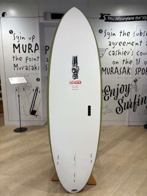 IMG_8814 - SHOP NEWS | ムラサキスポーツオンラインストア 通販 White surfboard with a green edge on a wooden display rack inside a shop, graffiti-style walls in the background.