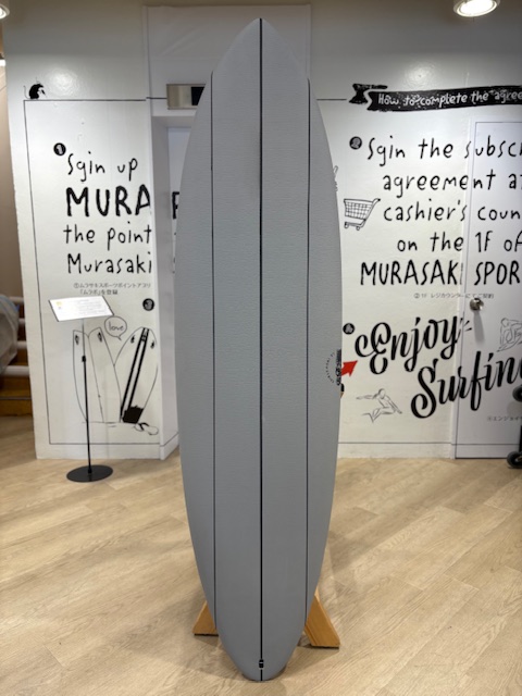 IMG_8815 - SHOP NEWS | ムラサキスポーツオンラインストア 通販 Grey surfboard standing upright on a wooden display rack inside a shop or showroom, with bold wall graphics in the background.