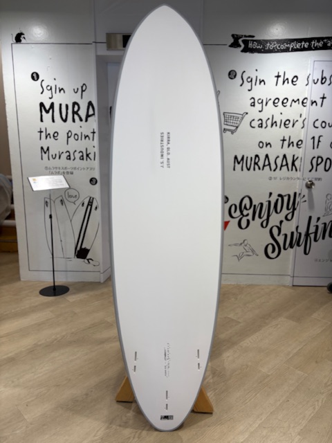 IMG_8816 - SHOP NEWS | ムラサキスポーツオンラインストア 通販 White surfboard standing upright on a wooden display rack in a showroom, with graffiti-style text on the walls behind.