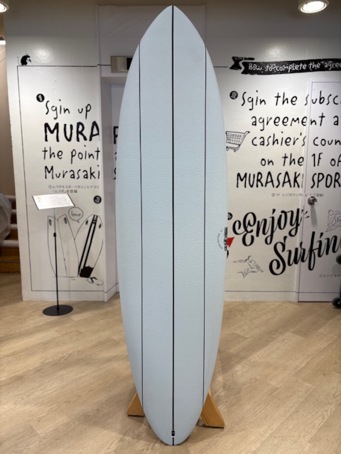 IMG_8817 - SHOP NEWS | ムラサキスポーツオンラインストア 通販 Light blue surfboard on a wooden stand indoors, centered between wall murals and showroom signs.