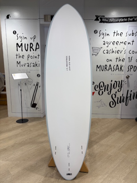 IMG_8818 - SHOP NEWS | ムラサキスポーツオンラインストア 通販 White surfboard on a wooden stand in an indoor exhibition, with bold, graffiti-style text on the walls behind.