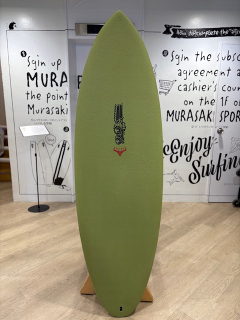IMG_8821 - SHOP NEWS | ムラサキスポーツオンラインストア 通販 Green surfboard standing upright on a wooden stand indoors, with a black logo centered and graffiti-style text on white walls in the background.