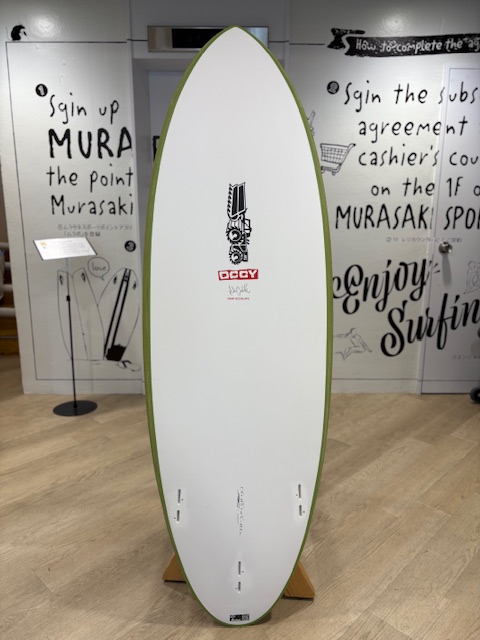 IMG_8822 - SHOP NEWS | ムラサキスポーツオンラインストア 通販 White surfboard with a lime-green edge on a wooden stand in a showroom, centered with a small black-and-white logo near the middle, and decorative wall text art in the background.