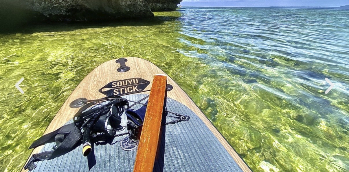 Stand-up paddleboard with gear on deck, cruising over clear emerald water near a rocky shoreline under bright sun.