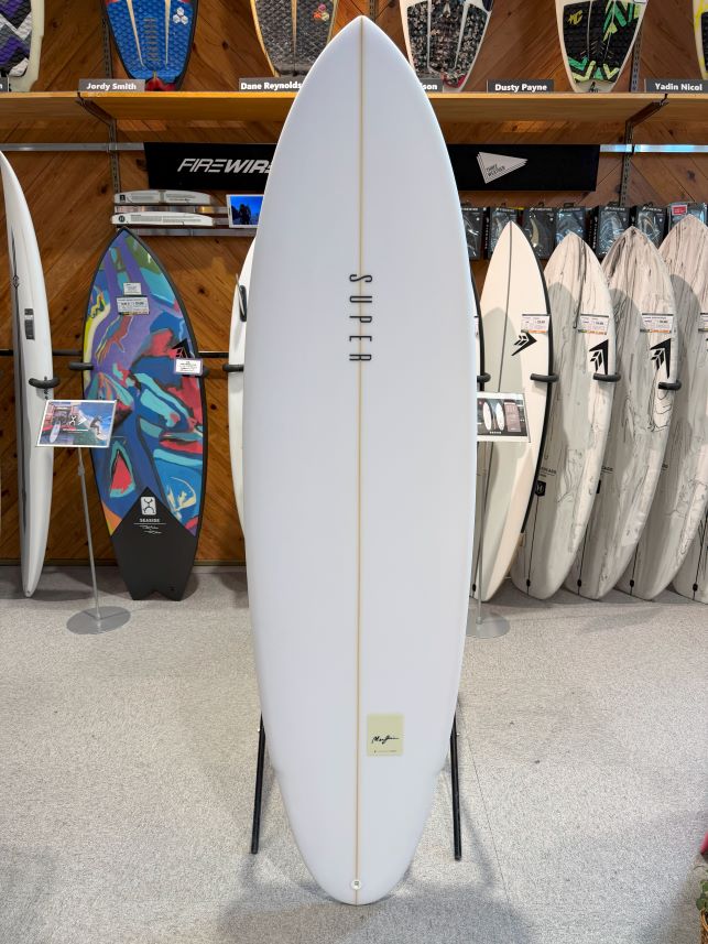 IMG_0483 - SHOP NEWS | ムラサキスポーツオンラインストア 通販 White surfboard displayed upright on a stand in a surf shop, with other boards on racks in the background.