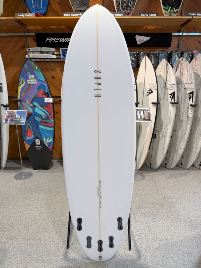 IMG_0484 - SHOP NEWS | ムラサキスポーツオンラインストア 通販 White surfboard on a display stand in a shop, centered with a vertical gold stringer and the word 'SUPER' running along its length, surrounded by other boards on racks.