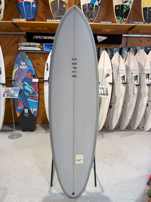 IMG_0486 - SHOP NEWS | ムラサキスポーツオンラインストア 通販 Large gray surfboard on a display stand in a shop, with racks of colorful boards in the background and wood shelving behind it.