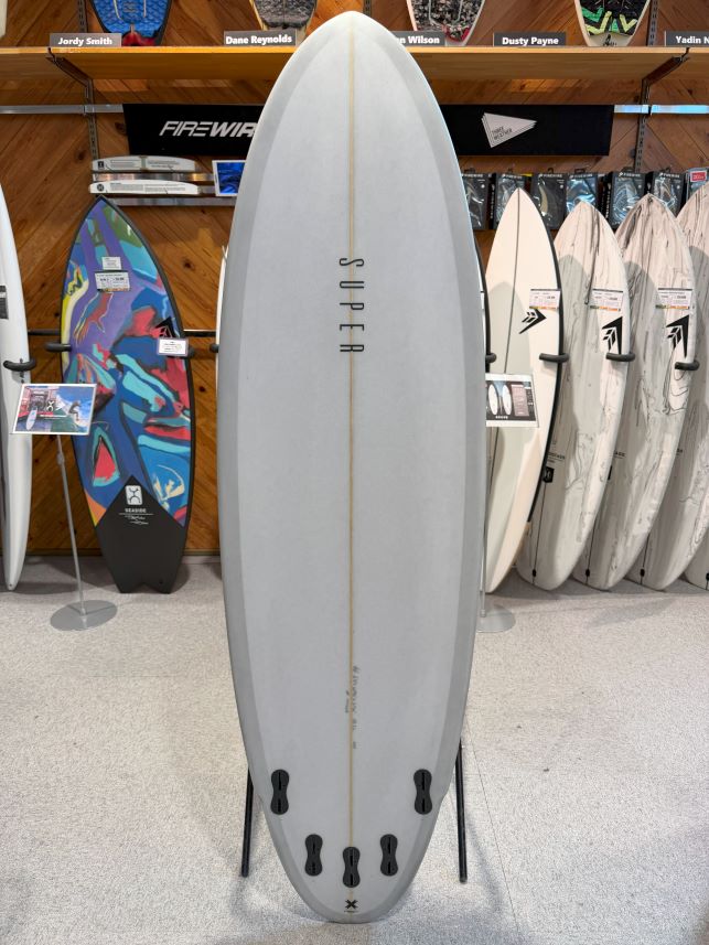 IMG_0487 - SHOP NEWS | ムラサキスポーツオンラインストア 通販 Light gray surfboard on a display stand in a shop, with other boards on shelves behind it.