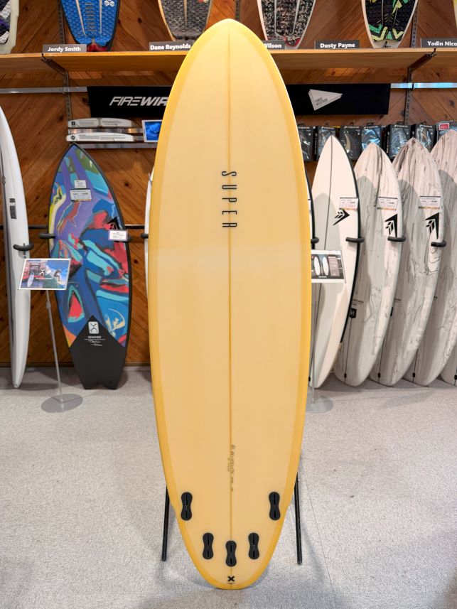 IMG_0491 - SHOP NEWS | ムラサキスポーツオンラインストア 通販 Yellow surfboard on display stand in a shop, surrounded by other boards and rack signage in the background.