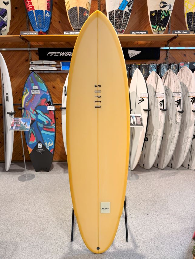 IMG_0492 - SHOP NEWS | ムラサキスポーツオンラインストア 通販 Yellow surfboard on a stand in a surf shop, surrounded by other boards on racks behind it.