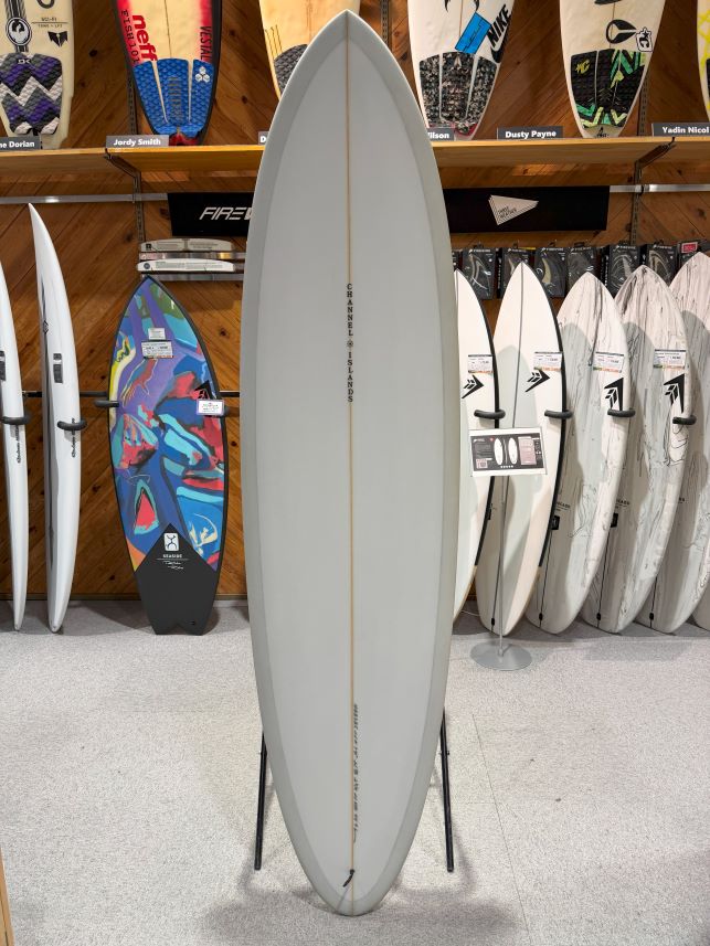 IMG_0564 - SHOP NEWS | ムラサキスポーツオンラインストア 通販 Tall light-gray surfboard on a stand in a surf shop, with colorful boards and racks in the background.