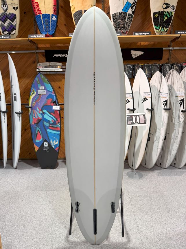 IMG_0565 - SHOP NEWS | ムラサキスポーツオンラインストア 通販 Tall light-gray surfboard on a display stand in a surf shop, with a single wood stringer and 'Channel Islands' printed along the center, surrounded by other boards on racks.