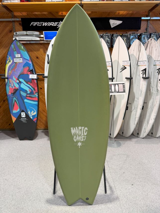 Olive green surfboard on display in a shop, with other boards in the background and a 'Magic' logo near the tail.