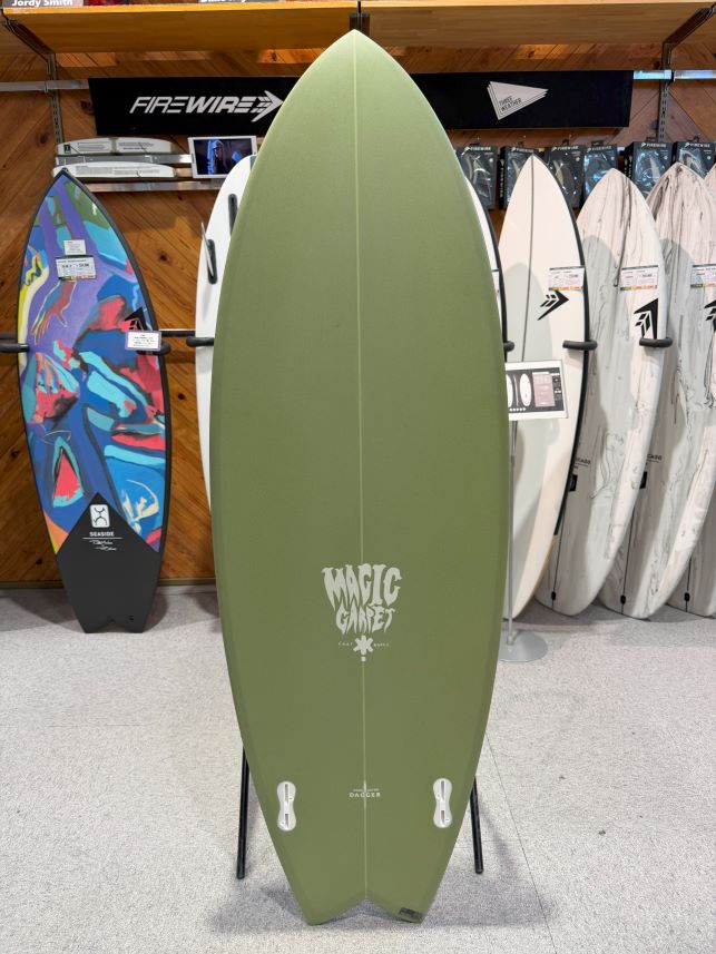 Olive green surfboard on a stand in a shop, featuring a 'Magic Carpet' logo near the bottom and two fin boxes, with other boards lined up behind.