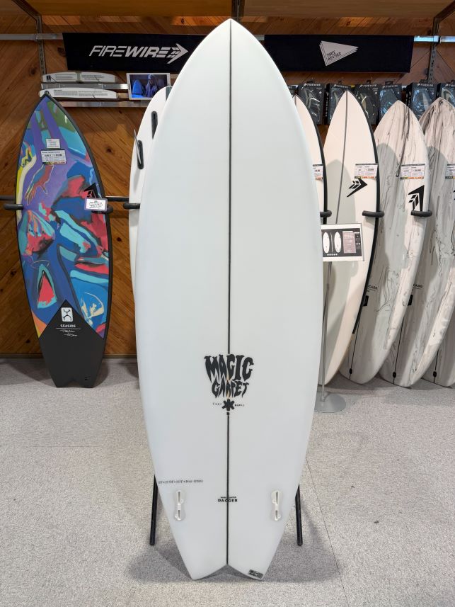 White surfboard on a display stand in a surf shop, with a 'Magic City' logo near the tail and other boards lined up in the background.