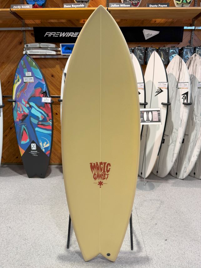Beige surfboard on a stand with a 'Magic Carpet' logo, displayed in a surf shop with other boards in the background.