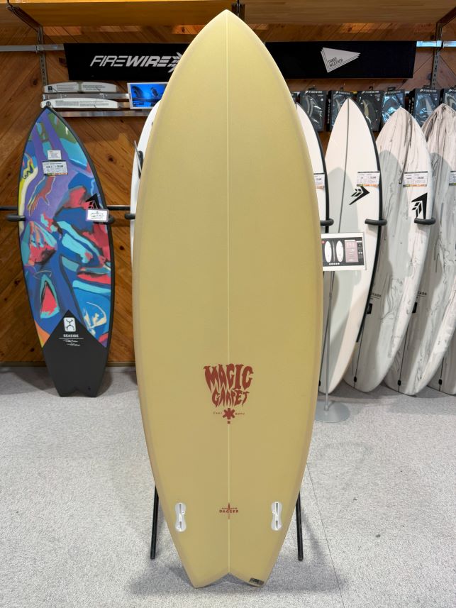 Beige surfboard on a display stand in a shop, with a red 'Magic Carpet' logo near the bottom, surrounded by other boards.