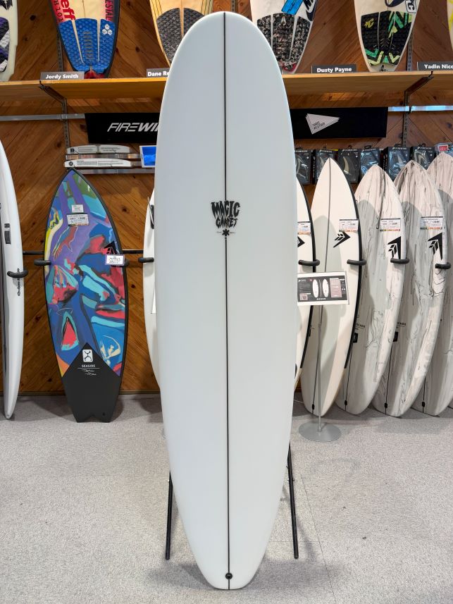 White surfboard on a stand in a surf shop, centered with a black logo reading 'Magic Carpet'.