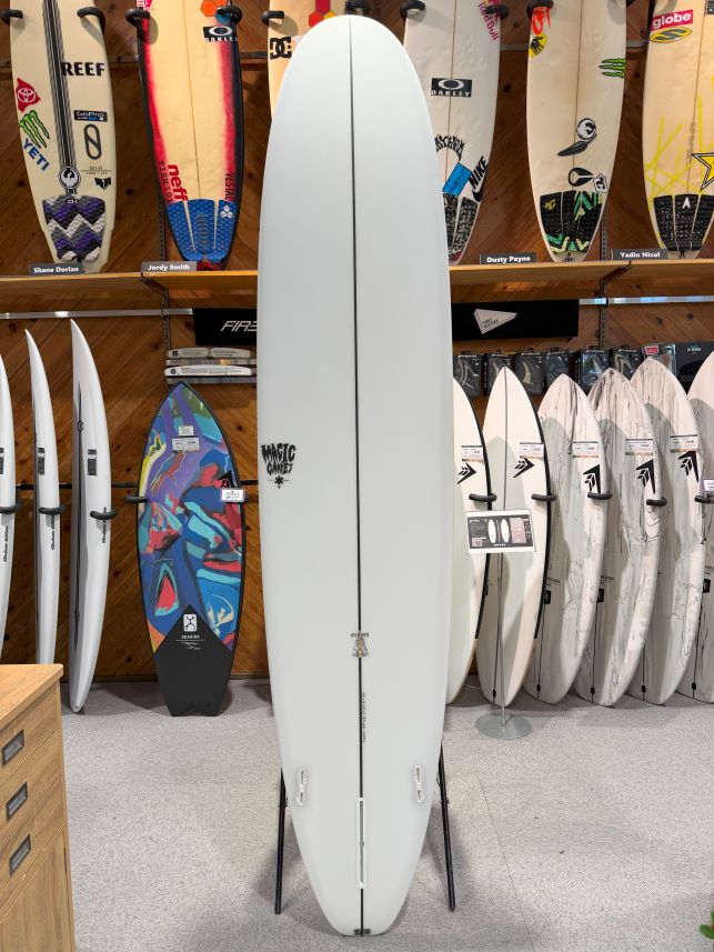 White surfboard displayed upright on a stand in a surf shop, with colorful boards on shelves behind it.