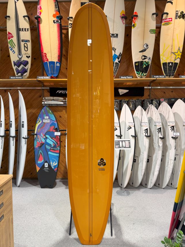 IMG_0622 - SHOP NEWS | ムラサキスポーツオンラインストア 通販 Orange longboard surfboard on a stand in a surf shop, surrounded by racks of white and colorful boards against a wooden wall shelves.