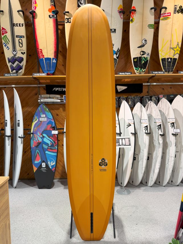 IMG_0623 - SHOP NEWS | ムラサキスポーツオンラインストア 通販 Orange longboard surfboard on a stand in a surf shop showroom, with other boards visible on walls around it.