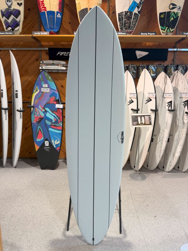 IMG_0662 - SHOP NEWS | ムラサキスポーツオンラインストア 通販 Large light-blue surfboard on a stand in a surf shop, with other boards on racks and wall behind.