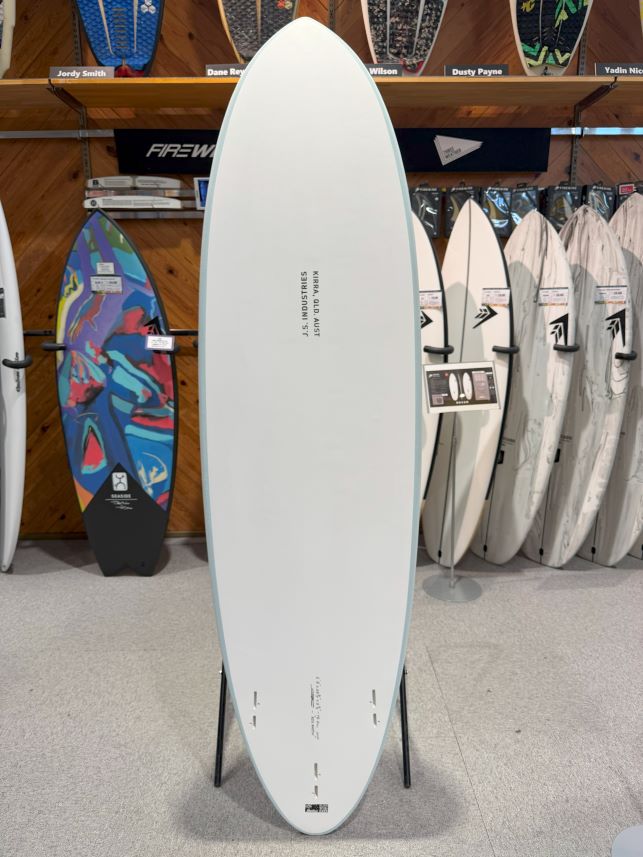IMG_0663 - SHOP NEWS | ムラサキスポーツオンラインストア 通販 White surfboard on a display stand in a surf shop, with colorful boards on racks behind it.