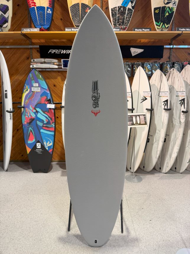 IMG_0666 - SHOP NEWS | ムラサキスポーツオンラインストア 通販 Gray surfboard on a stand in a showroom with other boards on display behind it, featuring a black logo and red emblem in the center.