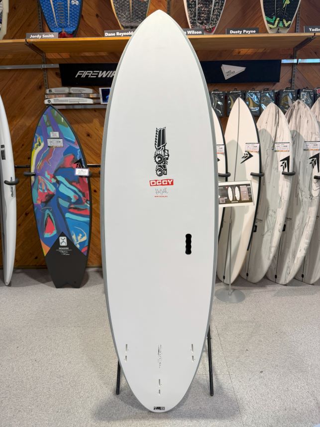 IMG_0667 - SHOP NEWS | ムラサキスポーツオンラインストア 通販 White surfboard on a display rack in a surf shop, with a black logo near the center and a stand at the bottom edges.
