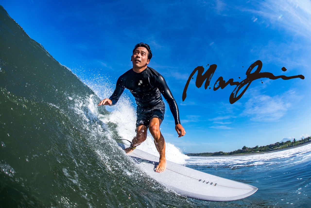 image0 2 - SHOP NEWS | ムラサキスポーツオンラインストア 通販 A man in a black wetsuit rides a white surfboard along a breaking wave under a bright blue sky.